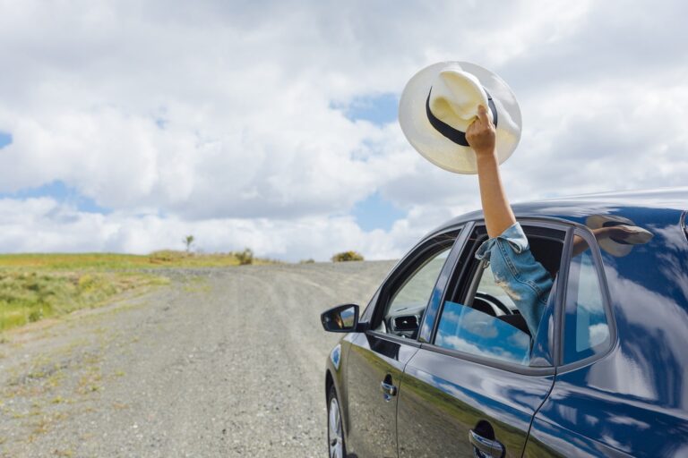 Blue car driving down the road while the woman in the back holds a hat out the window